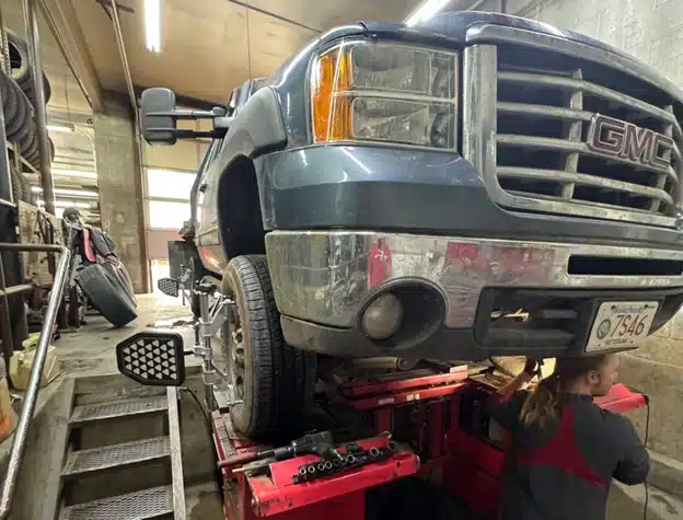 Fall is the perfect time for a wheel alignment check in Durango, CO with Firestone of Durango. Image of blue Chevy GMC pickup truck hooked up to the alignment machine in the shop with the mechanic performing checks underneath the truck.