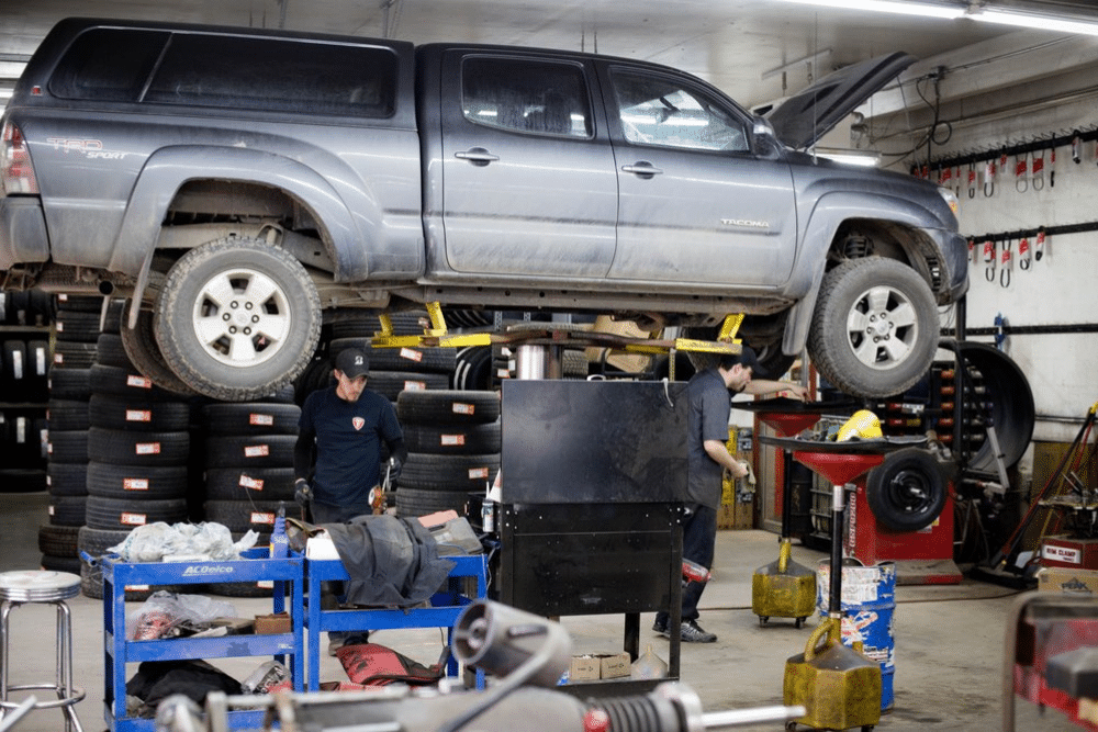 car maintenance tips, auto repair in Durango, CO at Firestone of Durango. Toyota Tacoma lifted on a hydraulic lift in an auto repair shop, with mechanics working underneath and tools scattered around.