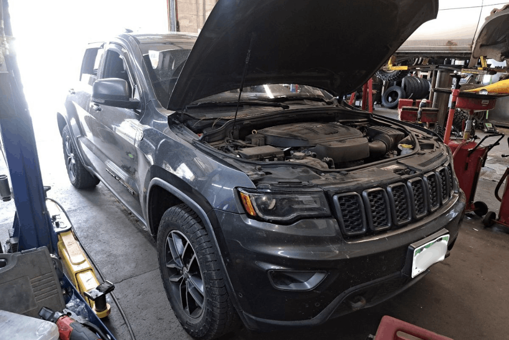 Car check, auto repair in Durango, CO at Firestone of Durango. Image of a gray Jeep Grand Cherokee inside an auto repair shop with the hood open, showing the engine bay during inspection and service.