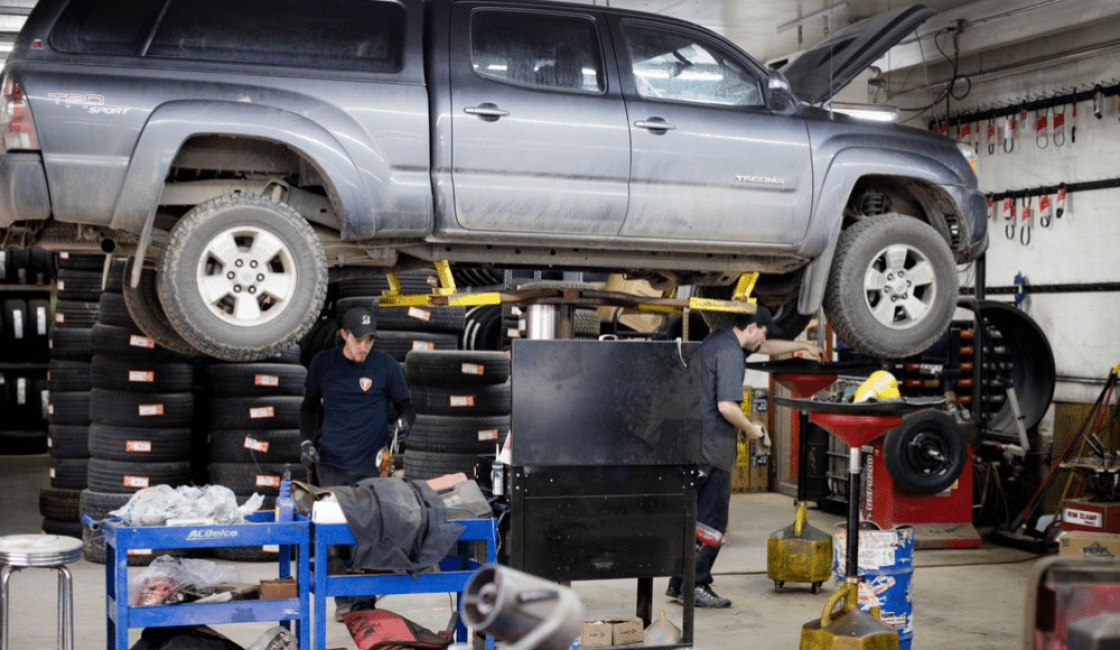 car maintenance tips, auto repair in Durango, CO at Firestone of Durango. Toyota Tacoma lifted on a hydraulic lift in an auto repair shop, with mechanics working underneath and tools scattered around.